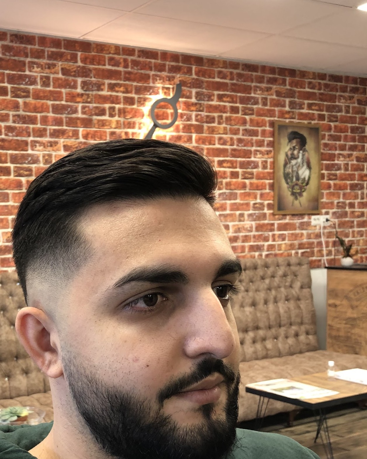 Klant met een strakke fade en een verzorgde baard in de zaak, met de bakstenen muur en een barbiersportret op de achtergrond