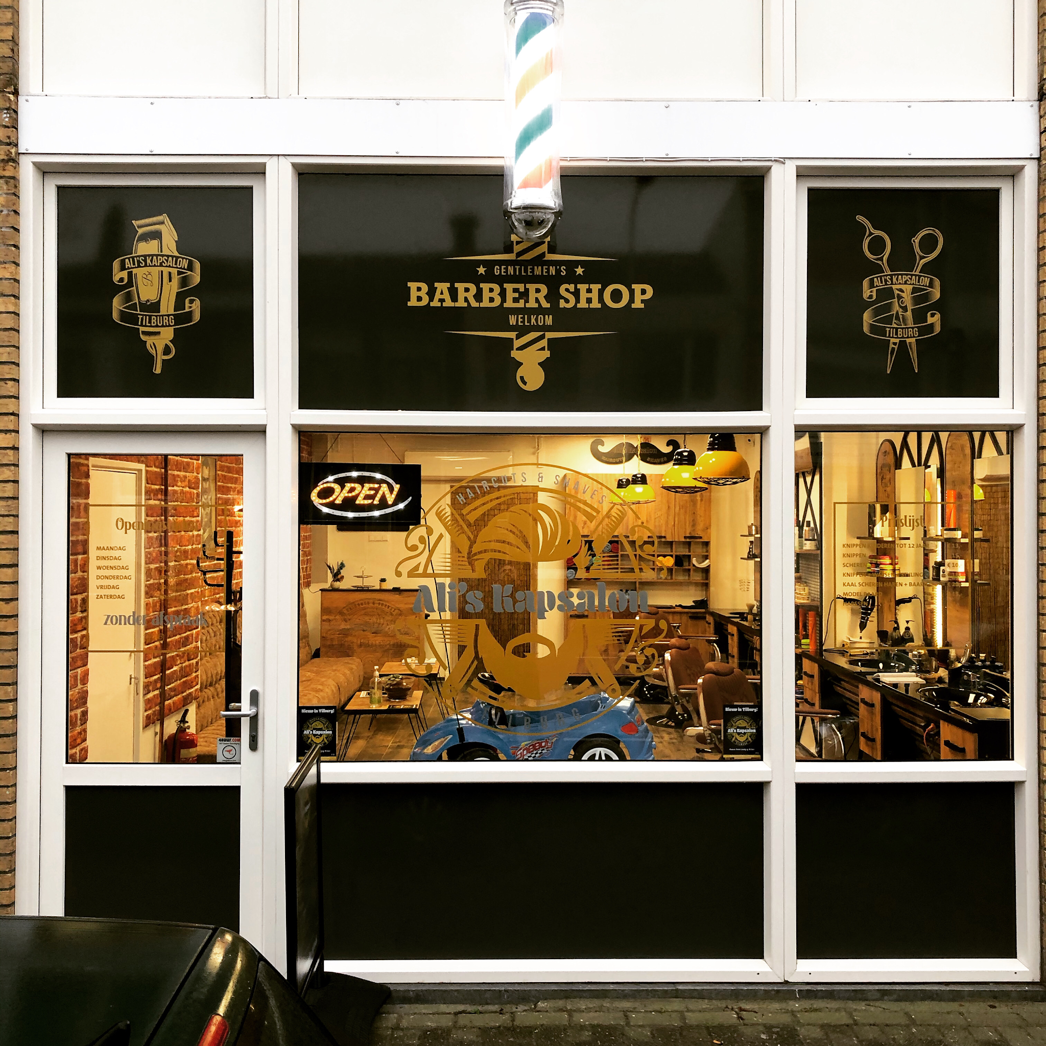 De etalage van Ali’s kapsalon aan de Besterdring: gouden belettering Gentlemen’s Barber Shop op zwart glas, een barbierspaal en een verlicht Open-bord